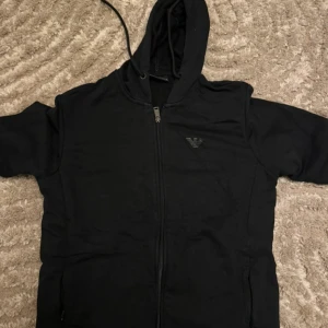 Svart hoodie från Armani med dragkedja - Svart hoodie från Armani med hel dragkedja framtill och klassisk huva. Tröjan har logga på bröstet och är tillverkad i mjuk bomullsmix. Perfekt för en chill och stilren look. Använd några gånger nypris: 1500kr