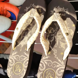 Bruna sandaler från Koala Bay med änglamotiv - Snygga bruna sandaler från Koala Bay med krämvita virkade remmar och unikt änglamotiv samt texten 'When Angels Sing' på sulan. Sulan har dekorativa mönster i beige och guld, och undersidan är präglad med Koala Bay-loggan. Kanonskick. Inga anmärkningar. 