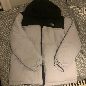 Grå och svart pufferjacka The North Face - Säljer en grå och svart pufferjacka i storlek XL (EU) Jackan har huva, dragkedja och är tillverkad i 100% polyester. Den är lätt, vindtät och vattenavvisande – perfekt för kalla dagar. Klassisk logga broderad på bröstet. Dragkedjan har gått av.
