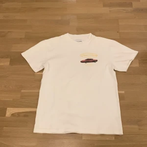 Vit Grand Fierens t-shirt med biltryck - Vit t-shirt från Grand Fierens by Braydon Alexander, tillverkad i 100% bomull. Framsidan har ett tryck med en röd sportbil och logga, medan baksidan har en stor bild av bilen ovanifrån och texten 'Tour' i gult. Skön passform och coolt bilmotiv.