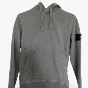 Grå hoodie från Stone Island - Säljer en grå hoodie från Stone Island med klassisk logga på ärmen. Tröjan har huva med snörning och en stor magficka framtill. Tillverkad i mjuk bomull och har en clean, stilren look som passar perfekt till streetwear äkta!!.