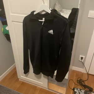 Svart Adidas hoodie med dragkedja - Svart hoodie från Adidas med vit logga på bröstet. Tröjan har dragkedja framtill, huva och långa ärmar. Tillverkad i mjukt och lätt polyester-material, perfekt för träning eller chill. Enkel och stilren design som funkar till det mesta.