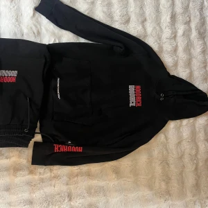 Svart hoodie från Hoodrich - Svart hoodie från Hoodrich med tryckt logga i vitt och rött på bröstet och ärmen. Tröjan har huva med snörning och en stor magficka framtill. Perfekt för en streetwear-look och chill dagar.