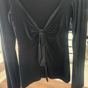 Svart långärmad topp med knyt - Svart långärmad topp från DesignBySi med djup v-ringning och snygg knyt framtill. Modellen är slim och har ett mjukt, stretchigt material. Använt den 1 gång.