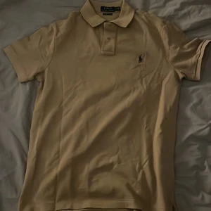 Beige pikétröja Polo Ralph Lauren M - Snygg beige pikétröja från Polo Ralph Lauren i modellen Custom Slim Fit. Klassisk krage, korta ärmar och ikonisk broderad logga på bröstet. Tillverkad i bomull för en skön och luftig känsla. Perfekt för en clean och stilren look. 