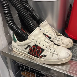 Gucci Ace sneakers med orm brodyr - Gucci Ace sneakers i vitt skinn med ikonisk röd och svart orm brodyr på sidan samt gröna och röda detaljer. Klassisk låg modell med rund tå och platt sula. Snygg streetstil med lyxig känsla och unika detaljer. Strl 46 Box och kvitto finns ÄKTA! Välanvända men i snyggt skick 