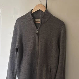 Grå stickad kofta från Sfera Men - Mörkgrå stickad kofta från Sfera Men med hög krage och dragkedja framtill. Regular fit och ribbade muddar vid ärmslut och nederkant. Perfekt för kyliga dagar och enkel att matcha med andra plagg.