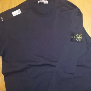 Svart långärmad tröja Stone Island - Svart långärmad tröja från Stone Island med klassisk rund hals och ikonisk logotyp-patch på vänster ärm. Tröjan är i mjuk bomull och har en stilren, enkel design som passar perfekt till jeans eller cargo. Märkeslappen sitter kvar.