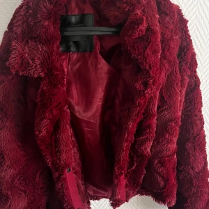 Vinröd pälsjacka från KappAhl - Mysig vinröd pälsjacka med fluffig känsla och snygg struktur i materialet. Jackan har en klassisk krage och stängs med knappar framtill. Perfekt för dig som vill sticka ut med en trendig look och hålla dig varm under kyliga dagar.• använd men inget som syns • fin jacka för sen vår eller tidig höst, men jag skulle också kunna säga att det skulle passa som en kofta till vintern 