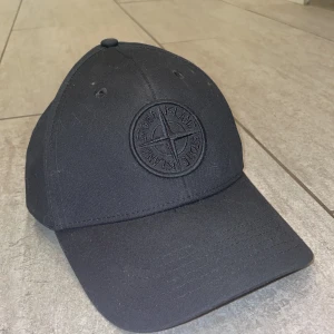 Svart keps från Stone Island - En sprillans ny svart keps från Stone Island med broderad logotyp framtill. Klassisk böjd skärm och justerbar rem baktill för perfekt passform. Tillverkad i bomull för en bekväm känsla och stilren look. Perfekt accessoar för att lyfta din outfit.