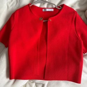 Röd bolerotopp från Zara - Säljer en röd bolerotopp från Zara med korta ärmar och rund halsringning. Toppen har en snygg guldig knapp framtill och är croppad i modellen. Materialet är mjukt och känns lite stickat, perfekt för att lyfta vilken outfit som helst. 