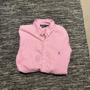 Rosa rutig skjorta Polo Ralph Lauren - Snygg rosa och vit rutig skjorta från Polo Ralph Lauren i slim fit. Skjortan har klassisk krage, knappar framtill och den ikoniska blå loggan broderad på bröstet. Perfekt för dig som gillar en fräsch och stilren look.