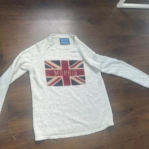 Vit stickad tröja Morris Co S - Vit stickad tröja från Morris Co i storlek Small. Framsidan har en stor Union Jack-flagga i rött, beige och mörkblått med texten 'Morris' i mitten. Tröjan har långärmad design och ribbad halsringning. Perfekt för dig som gillar klassisk stil med en twist.