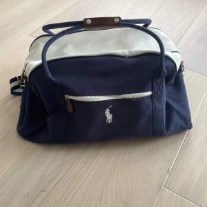 Blå och vit weekendbag från Polo Ralph Lauren - Snygg weekendbag från Polo Ralph Lauren i marinblått och vitt med klassisk logga framtill. Väskan har både handtag och justerbar axelrem, detaljer i brunt och dragkedja. Perfekt storlek för vardagen eller gym. Tillverkad i bomull och syntetmaterial.