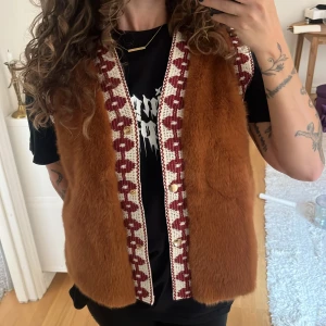 Brun pälsväst från Wild  - Säljer en brun pälsväst från Wild med mjuk fuskpäls och dekorativa virkade kanter i vinrött och vitt. Västen har knappar framtill och ett unikt, retroinspirerat mönster längs kanterna. Perfekt för dig som vill sticka ut med en cool och mysig look. En knapp saknas. Nypris 1200 sek