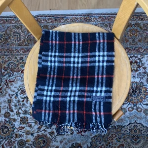 Rutig halsduk från Burberrys i ull - Klassisk rutig halsduk från Burberrys i 100% lammull. Färgerna är svart, vit och röd med fransar i ändarna. Perfekt accessoar för att lyfta din outfit med en tidlös brittisk känsla.
