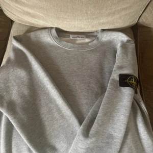 Grå sweatshirt från Stone Island - Snygg grå sweatshirt från Stone Island med klassisk rund hals och ikonisk logotyp-patch på ärmen. Tröjan är i mjuk bomull och har en clean look som passar perfekt till jeans eller joggers. Perfekt för dig som gillar streetwear och stilren design. Storlek M, typ aldrig använd.