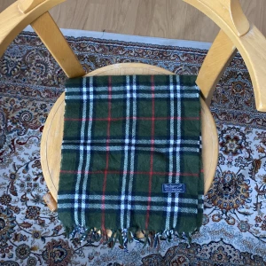 Grön rutig halsduk från Burberrys - Snygg grön halsduk från Burberrys i klassiskt rutigt mönster med inslag av vitt, svart och rött. Gjord i 100% lammull och har fransar i ändarna. Perfekt accessoar för att lyfta din outfit med en tidlös brittisk vibe.