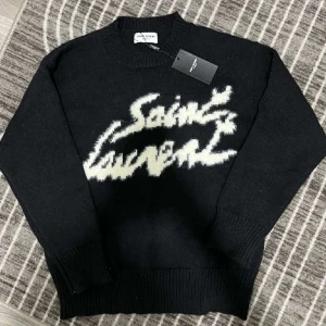 Svart stickad Saint Laurent tröja - Svart stickad tröja från Saint Laurent med vit, fuzzy logotyp framtill. Klassisk rund halsringning och ribbade muddar vid ärmslut och nederkant. Perfekt för dig som gillar exklusiva streetwear-vibbar och vill sticka ut med en ikonisk look.