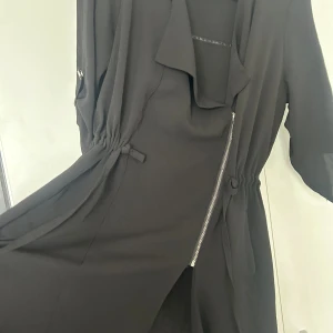 Svart lång kofta  - Svart lång kofta asymmetrisk dragkedja och knyt i midjan. Klänningen har trekvartsärmar med knappdetalj och en snygg, lös passform. Materialet känns lätt och flowy, perfekt för dig som gillar stilrena och moderna plagg.