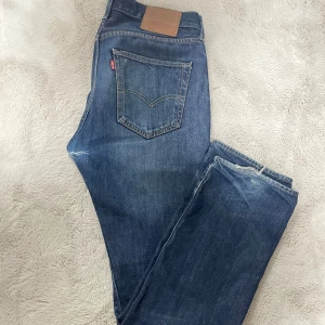 Levi's 501 blå jeans W31 L32 - Klassiska Levi's 501 jeans i blå denim med raka ben och normal passform. Jeansen har fem fickor, knappgylf och den ikoniska läderpatchen bak i midjan. Snyggt slitna detaljer och tydliga sömmar. Perfekta för en avslappnad och tidlös look. OBS! De har hål i skrevet men som enkelt kan lagas. Jag kan skicka med lag-patch + tråd i samma tvätt som jeansen. Frågor? Skriv!