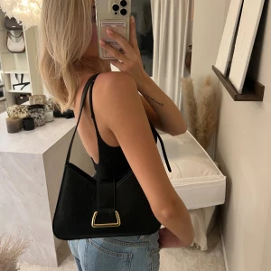Svart axelväska med gulddetalj - Stilren svart axelväska i skinnimitation med justerbart axelband och snygg guldfärgad spänndetalj framtill. Väskan har en triangulär form och stängs med dragkedja. Perfekt för dig som gillar minimalistisk och trendig design.