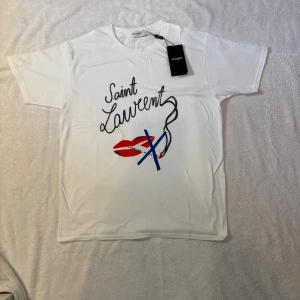 Vit Saint Laurent t-shirt med tryck - Otroligt snygg Vit t-shirt från Saint Laurent med coolt grafiskt tryck framtill som inkluderar röda läppar, blått kryss och svart text. Klassisk passform och rund hals. Storlek M . Tillverkad i mjuk bomull, perfekt för en clean och trendig look. Modellen är 179 65kg.