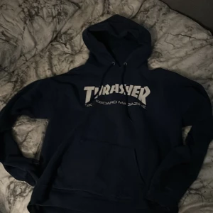 Mörkblå Thrasher hoodie med tryck - Säljer en mörkblå hoodie från Thrasher med klassiskt vitt logotryck på bröstet. Tröjan har huva med snörning och en stor magficka. Perfekt för dig som gillar streetwear och skatekänsla. Men den är lite sliten