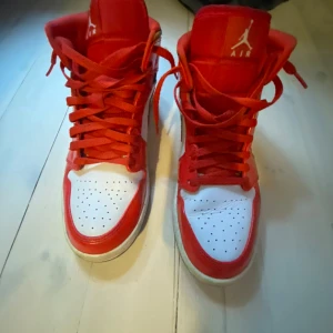 Nike Air Jordan 1 High röd/vit - Säljer ett par Nike Air Jordan 1 High i rött och vitt med klassisk swoosh på sidan. Skorna har snörning, hög siluett och genomskinlig sula. Tillverkade i skinn med färgglada detaljer vid ankeln. Perfekta för dig som vill sticka ut med en ikonisk sneaker.