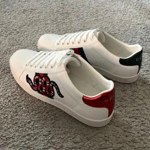 Gucci Ace sneakers med orm  - Gucci Ace sneakers i vitt skinn med klassiska röd-gröna ränder på sidorna och broderad orm på ena skon. Baktill har ena skon röd metallic-detalj och den andra grön, båda med Gucci-logga. Snygg och lyxig modell med snörning och platt sula. Vid frågor kontakta 