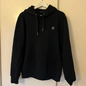 Svart hoodie från Morris, storlek M - Svart hoodie från Morris med dragsko i huvan och liten broderad logga på bröstet. Klassisk modell med ribbade muddar och mjukt material som känns skönt mot huden. Perfekt för en chill och stilren look.