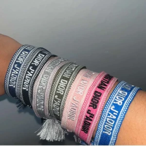 Färgglada armband - Säljer flera  armband i olika färger som blå, rosa, grå, svart och olivgrön. Armbanden är vävda i textil med broderad logga och har tofsar i ändarna. Perfekt accessoar för att lyfta din outfit med en lyxig touch.