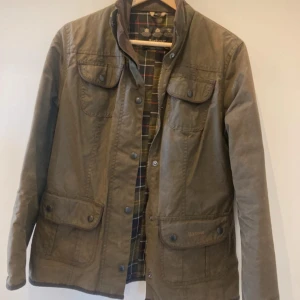 Olivgrön Barbour jacka med rutigt foder - Klassisk olivgrön jacka från Barbour med rutigt innerfoder, fyra stora fickor med tryckknappar och hög krage med brun manchester. Jackan har dragkedja och knappar framtill, perfekt för höst och vår. Snygg och tidlös design som funkar till allt.