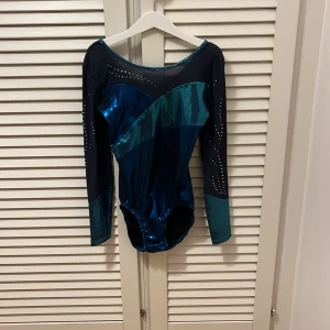 Blå, turkos glittrig gymnastikdräkt  - Säljer en långärmad gymnastikdräkt i glansigt blå, turkos och marinblå med  stenar på ärmar och överdel och baksida. 