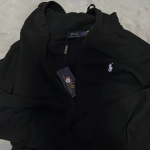 Svart hoodie från Polo Ralph Lauren XL - Svart hoodie från Polo Ralph Lauren i storlek XL. Klassisk design med huva, dragkedja och den ikoniska vita Polo-loggan broderad på bröstet. Perfekt för en chill och stilren look. Mjuk och skön bomullskvalitet som passar till alla tillfällen. Passar också L.