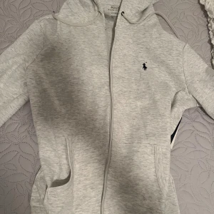 Ljusgrå Polo Ralph Lauren Kofta/Hoodie - Ljusgrå hoodie från Polo Ralph Lauren med Tröjan har luva med snörning och två fickor framtill. Den är ganska stor i storleken så den passar M.