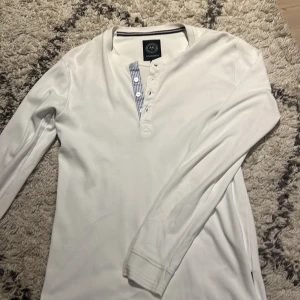 Vit långärmad tröja från Lindbergh - Vit långärmad tröja från Lindbergh med knappar framtill och en snygg detalj i form av blå- och vitrandigt tyg vid knappslån. Tröjan har rund halsringning och är tillverkad i mjuk bomull, perfekt för en clean och stilren look. Kom gärna med prisförslag!!!