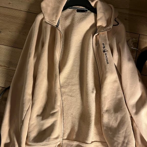 Beige hoodie från Sail Racing med huva - Beige hoodie från Sail Racing med dragkedja och broderad logga på bröstet. Mjuk fleece på insidan, huva och praktisk bröstficka med dragkedja. Perfekt för kyliga dagar och snygg att slänga på sig över en t-shirt.