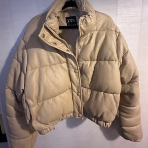Beige croppad pufferjacka från Zara - Säljer en beige croppad pufferjacka från Zara med quiltad design och hög krage. Jackan har tryckknappar framtill och är tillverkad i syntetmaterial med skinnliknande yta. Perfekt för dig som gillar trendiga och oversized silhuetter.