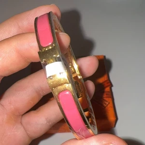 Harmens Click H armband mörkrosa  - Snyggt armband från Harmans i guldtonad metall med blanka rosa emaljdetaljer. Klassisk oval form och diskret logga på insidan. Perfekt accessoar för att lyfta din outfit med en färgklick och lyxig känsla. Kommer i box, är rostfritt och vattentåligt. 