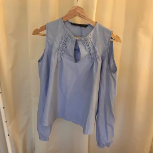 Ljusblå blus med cut out från Zara - Supersöt ljusblå blus från Zara Trafaluc Collection med långa ärmar och coola cut out-detaljer vid axlarna. Framsidan har rynkade detaljer och en liten knapp vid halsen. Perfekt för dig som gillar unika detaljer och en luftig känsla.