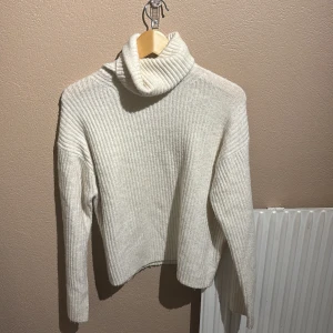 Vit ribbad polotröja i ullmix - Mysig vit/ lite beige polotröja med ribbad struktur och hög krage. Tröjan är stickad i en mjuk ullmix och har långärmad passform. Perfekt för kyliga dagar när du vill vara både varm och stilren.