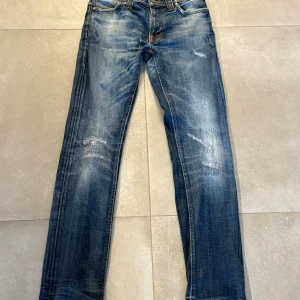 Blå jeans från Nudie Jeans Co - Snygga blå jeans från Nudie Jeans Co med slitna detaljer och klassisk femficksdesign. Jeansen har orangea sömmar, normal passform och raka ben. Tillverkade i bomull med en cool tvättad look och tydliga slitningar på låren och knäna.