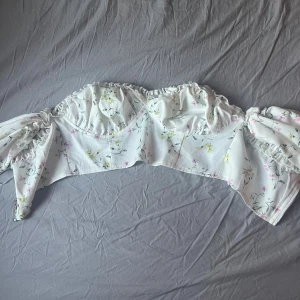 Vit blommig korsett top från Missguided - Superfin vit korsett top från Missguided med blommigt mönster i ljusgult och rosa. Toppen har volangdetaljer längs kanten och är axelbandslös med en snygg croppad passform. Materialet är mjukt och lätt, perfekt för varma dagar. Säljer pga ingen användning!