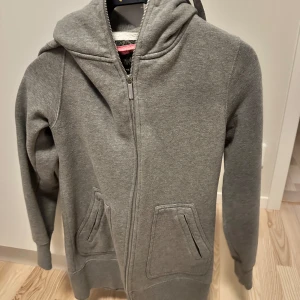Grå hoodie från Warp med dragkedja - Mysig grå hoodie från Warp med hel dragkedja och två fickor framtill. Tröjan har huva och är fodrad med mjuk fleece på insidan, perfekt för kyliga dagar. 