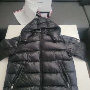 Svart Moncler dunjacka med huva - Säljer en svart dunjacka från Moncler med glansig finish, huva och två dragkedjefickor framtill. Jackan har klassisk Moncler-logga på ärmen och coola detaljer som quiltad design och innerfoder med serietidningsmönster. Perfekt för kalla dagar när du vill hålla stilen.