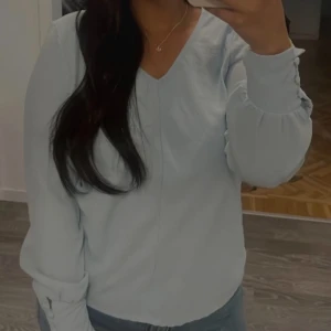 Ljusblå v-ringad blus - Snygg ljusblå blus med v-ringning och långa ärmar. Ärmarna har en lätt volangdetalj vid mudden som ger en extra touch. Perfekt att matcha med jeans eller kjol för en fräsch look.
