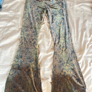 Mönstrade bootcut byxor Urban Outfitters - Otroligt snygga och bekväma bootcut byxor från Urban Outfitters med elastisk midja. Byxorna har ett unikt barockinspirerat mönster i guld och mörkgrön/brun ton, tillverkade i ett glansigt, mjukt sammets material. 