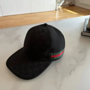 Gucci Keps - Snygg svart keps från Gucci med diskret GG-monogram över hela kepsen och klassiska gröna och röda ränder på sidan. Justerbar rem baktill i skinn och böjd skärm. Tillverkad i bomull och detaljer i skinn, perfekt för en trendig streetstyle Storlek L.
