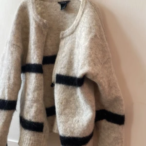 Beige kofta med svarta ränder från Lindex - Mysig beige kofta från Lindex med svarta, breda horisontella ränder. Koftan är stickad och har en enkel knäppning framtill. Perfekt för lager-på-lager och har en avslappnad, lite oversized passform med långa ärmar.